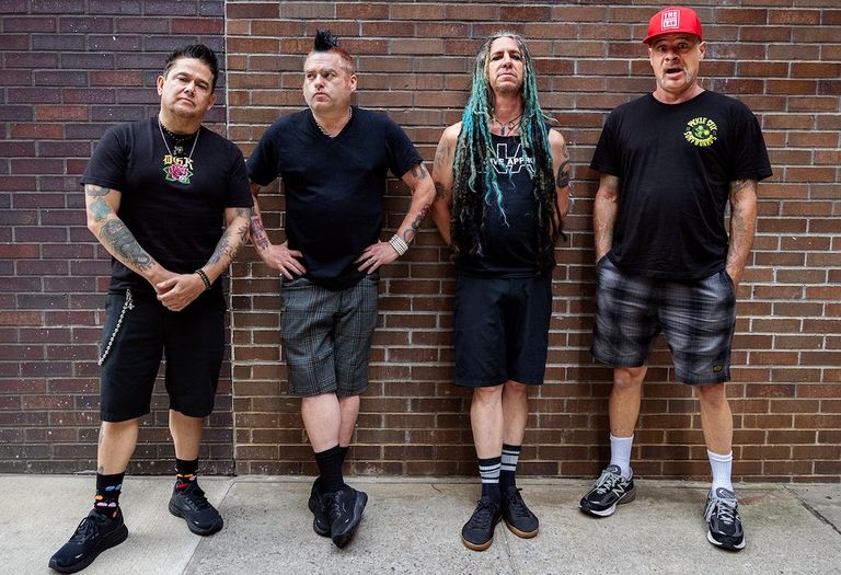 Promotional portrait of American punk rock band NOFX featuring Fat Mike and band members leaning against a brown brick wall.